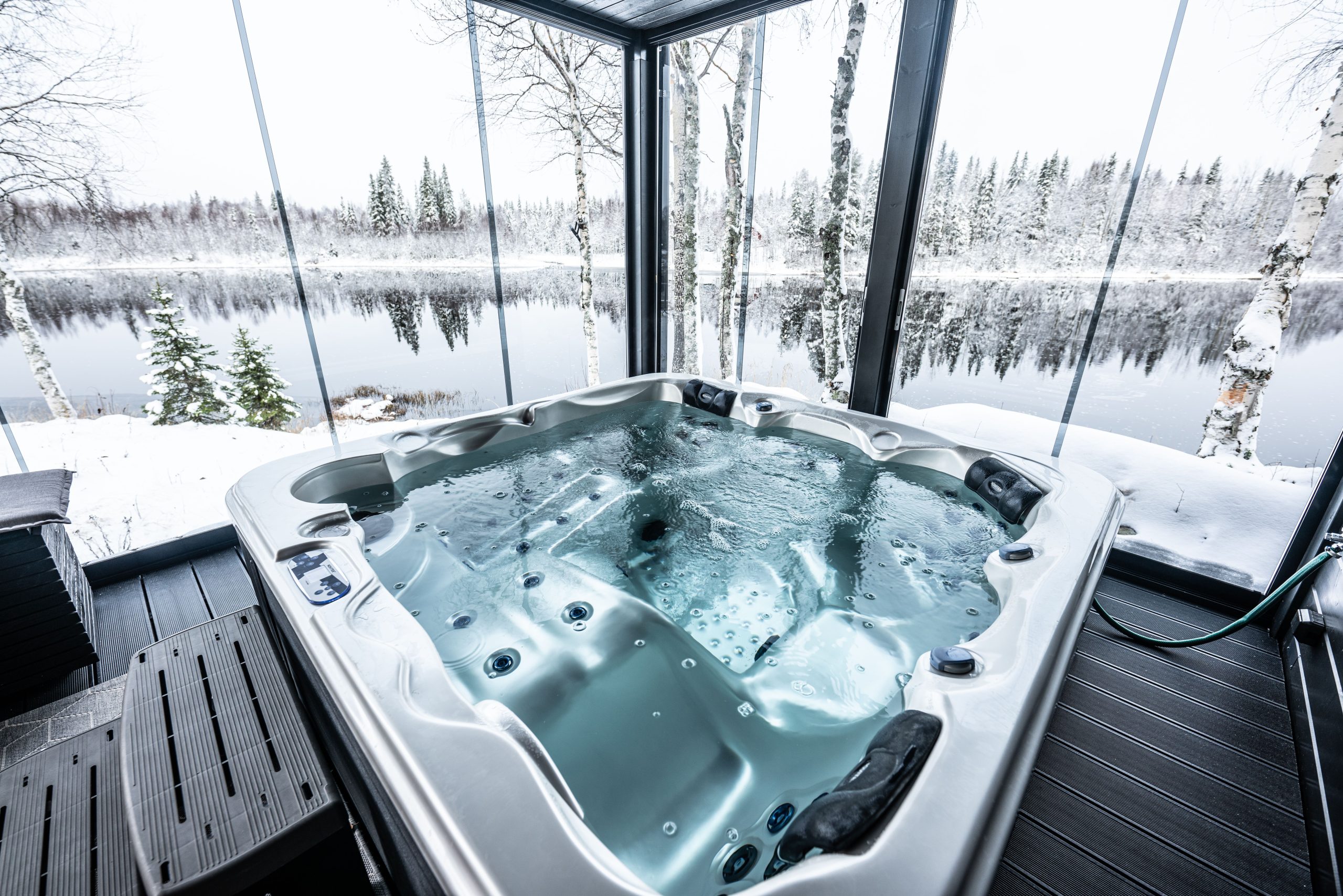 A luxurious hot tub on a glass-walled terrace, offering a panoramic view of a calm river and a snow-covered forest. The tub is filled with bubbling water, with snowy birch and spruce trees visible in the background.