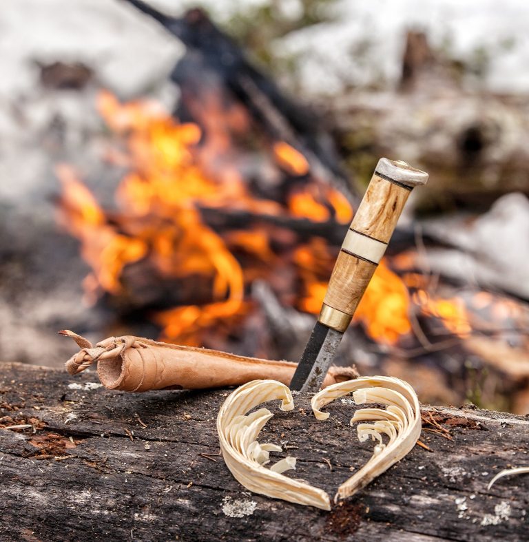 A knife with a wooden handle stands in a log beside a heart carved from wood shavings, with a campfire burning behind.